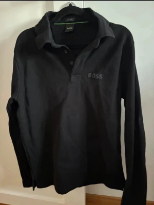 Svart långärmad polotopp från BOSS - Svart långärmad polotopp från BOSS med klassisk krage och knappar framtill. Regular fit och diskret BOSS-logga broderad på bröstet. Perfekt för en stilren och enkel look.