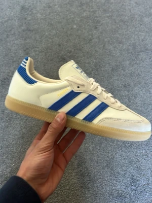 Adidas Samba OG vit/blå sneakers - Säljer ett par Adidas Samba OG sneakers i vitt skinn med blåa ränder och detaljer. Klassisk gummisula och guldfärgad Samba-text på sidan. Lågt skaft och retrovibe, perfekta till jeans eller shorts. Skön passform och tidlös design. Sprillans nya med box och allt 