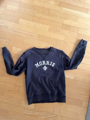 Blå sweatshirt från Morris - Mörkblå sweatshirt från Morris med vit logotyp och emblem på bröstet. Tröjan har rund halsringning, långa ärmar och ribbade muddar. Perfekt för en avslappnad och stilren look.