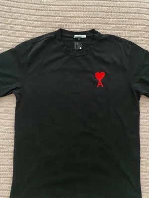 Svart t-shirt från Ami med rött hjärta - Svart t-shirt från Ami med klassisk passform och rund halsringning. På bröstet finns ett broderat rött hjärta med bokstaven A. Perfekt för en stilren och trendig look.