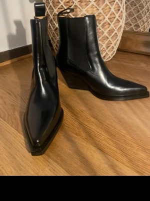 Svarta cowboy boots i skinn - Stilrena svarta cowboy boots med spetsig tå och snedklack. Tillverkade i blankt skinn med elastiska paneler på sidorna för enkel påtagning. Perfekta för dig som vill ha en edgy westernstil.