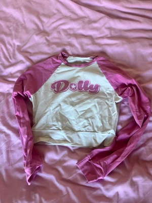 Vit och rosa långärmad Dolly-topp - Säljer en långärmad topp från Dolly style. Toppen har tryckt logga framtill och baktill. och är tillverkad i mjuk bomull. Obs. Har en liten fläck på ärmen (se bild) är även i barnstorlek men sitter som en xs 