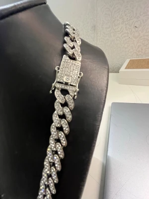 Silverfärgat cuban chain halsband - Stilrent halsband i cuban chain-modell med stora länkar, helt täckt av gnistrande stenar för en lyxig look. Silverfärgad metall och robust lås med stenar. Perfekt för dig som vill sticka ut med accessoarerna.