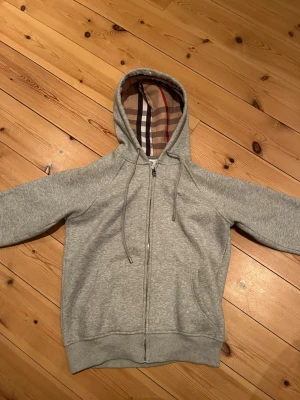 Grå zip-hoodie från Burberry - Snygg grå hoodie från Burberry med dragkedja och klassiskt rutigt foder i huvan. Mjukt material, dragsko i huvan och två fickor framtill. Perfekt för en chill och stilren look.