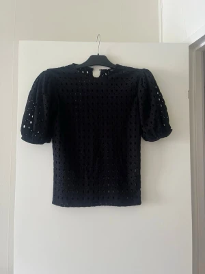 Svart blus med puffärm och broderade hål - Svart blus med korta puffärmar och broderade hål över hela tyget. Toppen har rund halsringning och knäppning bak i nacken. Perfekt för dig som gillar en luftig och trendig look.