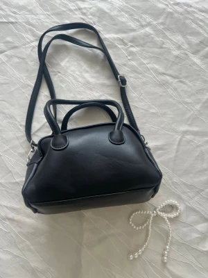 Svart handväska med axelrem - Stilren svart handväska i slät konstläder med både handtag och justerbar axelrem. Väskan har en mjuk, rundad form och silverfärgade detaljer. Perfekt för dig som gillar minimalistisk och klassisk design. Pris kan diskuteras!!