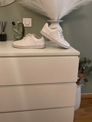 Vita Nike Air Force 1 sneakers - Klassiska vita Nike Air Force 1 sneakers med lågt skaft. Skorna har en stilren design med perforerade detaljer på tån och Nike-logga på sidan. Tillverkade i läder med gummisula för bra grepp. Perfekta för en clean och tidlös look.