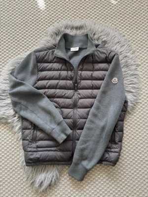 Grå cardigan från Moncler (M) - Säljer en snygg grå dunväst från Moncler med quiltad front och stickade ärmar. Västen har dragkedja, hög krage och Moncler-logga på ena ärmen. Perfekt för lager-på-lager och ger en cool look till hösten och vintern.