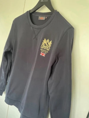 Mörkblå sweatshirt från Morris - Mörkblå sweatshirt från Morris med broderad guldfärgad logga och brittisk flagga på bröstet. Tröjan har rund halsringning och långa ärmar. Perfekt för en stilren och avslappnad look.