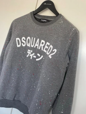 Grå DSQUARED2 sweatshirt med tryck - Grå sweatshirt från DSQUARED2 med vitt tryck och japanska tecken på bröstet. Tröjan har färgstänk i vitt och rött över hela ytan, rund halsringning och ribbade muddar. Perfekt för en trendig och avslappnad stil.