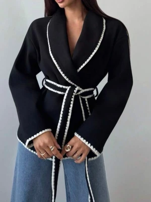 Svart cardiganjacka med vit kant - Stilren svart cardiganjacka med bred sjalkrage och vita dekorativa kanter. Jackan har ett matchande knytband i midjan och vida ärmar. Perfekt för dig som vill ha en trendig och bekväm look. Materialet ser ut att vara mjukt och lite tjockare, passar bra till jeans.