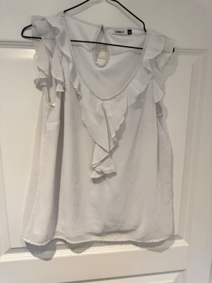 Vit volangtopp från ONLY - Säljer en söt vit topp från ONLY i storlek 36. Toppen är ärmlös med volangdetaljer längs axlar och ringning samt en liten öppning bak med knapp. Perfekt för dig som gillar en feminin och luftig stil.