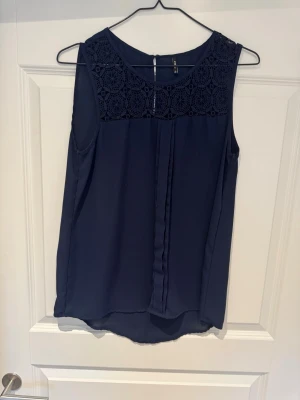 Mörkblå ärmlös topp från ONLY - Snygg mörkblå ärmlös topp från ONLY med spetsdetaljer över axlar och bröst. Toppen har rund halsringning och ett lätt, luftigt tyg som faller fint. Perfekt att styla med jeans eller kjol för en trendig look.
