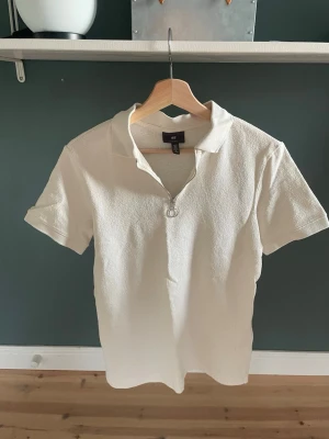 Vit pikétröja från H&M Regular Fit - Snygg vit pikétröja från H&M i regular fit. Tröjan har korta ärmar, klassisk krage och en knapp framtill. Tillverkad i ett strukturerat tyg som ger en modern känsla. Perfekt för en clean och stilren look. Endast testad
