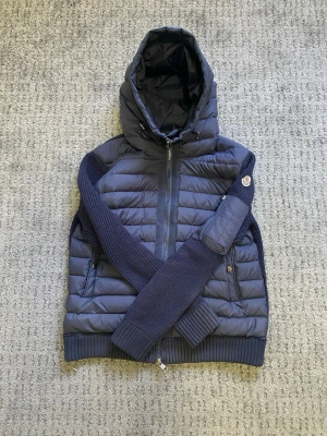 Mörkblå Moncler cardigan - Snygg mörkblå dunjacka från Moncler med stickade ärmar och huva. Jackan har dragkedja framtill, två fickor med knappar och ribbade muddar. Perfekt för kalla vinterdagar och har klassisk Moncler-logga på ärmen. Passar M!!