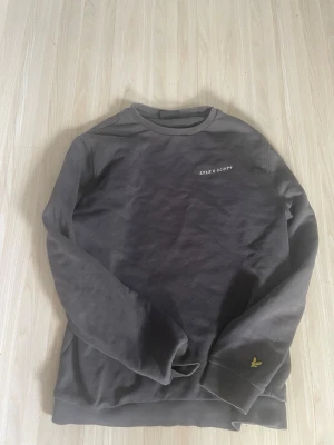 Mörkgrå sweatshirt från Lyle & Scott - Mörkgrå sweatshirt från Lyle & Scott med rund halsringning och diskret logga på bröstet. På ärmen finns den klassiska gula fågeln broderad. Tröjan har ribbade muddar vid ärmslut och nederkant, perfekt för en avslappnad stil.