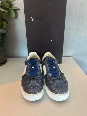 Valentino flycrews  - Snygga sneakers från Valentino med glittrig blå ovandel, vita detaljer på sidorna och vita sulor. Skorna har blå skosnören och en klassisk låg siluett. Perfekta för dig som vill sticka ut med stil och exklusiv känsla. Box medföljer, inte original 