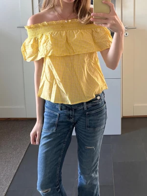 Gul off shoulder topp med volang - Säljer en söt gul off shoulder topp med volang och smockad överdel. Toppen har ett rutigt mönster i vitt och gult och är tillverkad i ett lätt bomullstyg. Perfekt för sommarens varma dagar och ger en fräsch och trendig look.