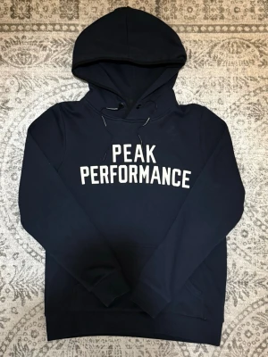 Mörkblå hoodie från Peak Performance - Snygg mörkblå hoodie från Peak Performance med vit logga tryckt på bröstet. Klassisk modell med huva och dragsko samt en stor magficka. Perfekt för en avslappnad och sportig stil.