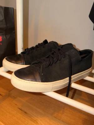 Axel Arigato  - Snygga svarta sneakers med ovandel i mocka och tåparti i läder. Skorna har svarta skosnören och en tjock vit gummisula som ger en clean look. Perfekta för dig som gillar minimalistisk stil och vill ha något som funkar till det mesta.