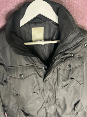 Field jacket från J.Lindeberg - Snygg pufferjacka från J.Lindeberg med hög krage och framfickor med lock och tryckknappar. Jackan har dragkedja och knappar framtill samt quiltad design. Perfekt för både kalla och även lite varmare dagar samt ger en stilren look. Skriv vid minsta funderingar😊