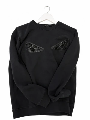Svart sweatshirt från Fendi med nitar - (OBS 2 bitar saknas på varje öga) Cool svart sweatshirt från Fendi med två ögonformade detaljer på bröstet, dekorerade med svarta nitar för en edgy look. Tröjan har rund halsringning och är tillverkad i mjukt bomullsmaterial. Perfekt för dig som vill sticka ut med en unik design.