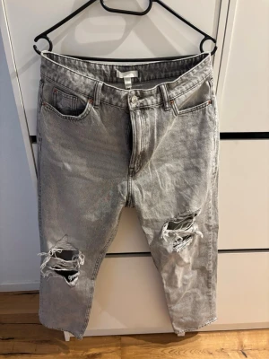 Grå slitna jeans från H&M - Grå jeans från H&M i storlek 42 med raka ben och coola slitningar på båda knäna. Klassisk femficksmodell och knappgylf. Perfekta för en avslappnad streetstyle-look.