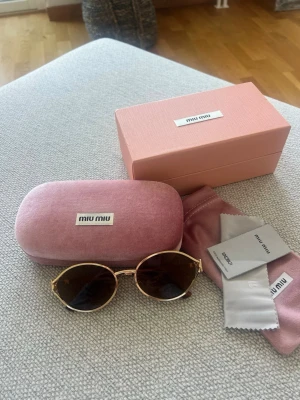 Miu Miu Solglasögon  - Exklusiva Miu Miu-solglasögon med eleganta guldfärgade metallbågar och bruntonade glas. Stilren och feminin modell med den ikoniska Miu Miu-loggan på sidorna som ger en lyxig touch.  Kommer som komplett set med originalbox, fodral, putsduk och booklet.  Skick: Mycket fint skick – sparsamt använda och väl omhändertagna.  Detaljer: • Märke: Miu Miu • Guldfärgade metallbågar • Bruntonade glas • Miu Miu-logga på sidorna • Komplett set med box, fodral och putsduk • Elegant och tidlös design