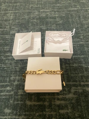 ÄKTA Guld Armband från Lacoste NYTT MED ALLA FÖRPACKNING - Snyggt guldigt armband från Lacoste med en ikonisk krokodil i mitten. Armbandet har en chunky kedjedesign och är tillverkat i metall med guldfinish. Levereras i original ask och med en vit tygpåse med Lacoste-logga. Perfekt accessoar för att lyfta din stil.  NYTT MED ALLA FÖRPACKNING.      ÄKTA
