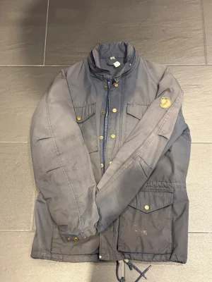 Mörkblå vintage vindjacka från Fjällräven - Snygg mörkblå vintage vindjacka från Fjällräven med klassisk logotyp på ärmen. Jackan har flera fickor med tryckknappar, hög krage och dragsko i nederkant. Perfekt för blåsiga dagar och enkel att matcha med olika outfits. Använd fåtal gånger men blivit smutsig, går att tvätta bort. Nypris 6000kr, säljer den för 1500kr men pris kan diskuteras pågrund av smuts osv. 