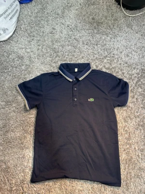 Mörkblå pikétröja från Lacoste - Snygg mörkblå pikétröja från Lacoste med klassisk krage och vita ränder på krage och ärmslut. Tröjan har korta ärmar, tre knappar framtill och den ikoniska gröna krokodilloggan broderad på bröstet. Perfekt för en stilren och sportig look.