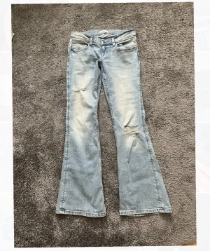 Ljusblå bootcut jeans från gina - Säljer ett par lågmidjade ljusblå bootcut jeans med slitningar och hål på ena benet från Gina Tricot. Näst intill helt oanvända, skriv om ni undrar nått som tex midjemått❤️