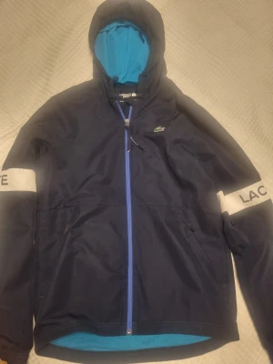 Marinblå Lacoste vindjacka med huva - Marinblå vindjacka från Lacoste Sport med blått meshfoder i huvan och dragkedja framtill. Jackan har vita detaljer på ärmarna med texten 'LACOSTE' och två fickor med dragkedja. Klassisk krokodillogga på bröstet och justerbar huva. Fast pris.
