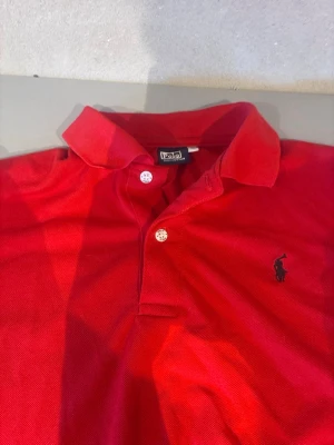 Röd pikétröja från Polo Ralph Lauren - Klassisk röd pikétröja från Polo Ralph Lauren med korta ärmar, krage och två knappar framtill. Tröjan har den ikoniska broderade loggan på bröstet och är tillverkad i bomull för en bekväm känsla.