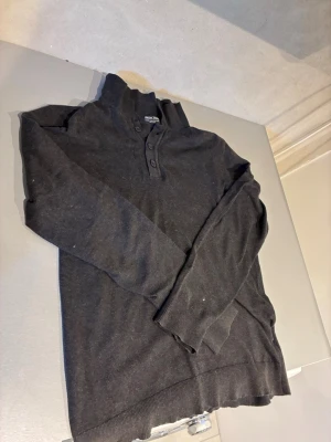 Mörkgrå tröja med knappar Indicode - Mörkgrå långärmad tröja från Indicode med hög krage och tre knappar framtill. Tröjan har ribbade muddar vid ärmslut och nederkant. Perfekt för en stilren och avslappnad look.