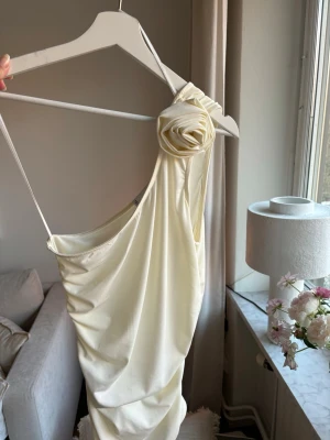 Krämvit oneshoulder topp med rosdetalj - Säljer en krämvit topp med oneshoulder och en snygg rosdetalj på axeln. Klänningen har smal axelrem och draperad passform som ger en cool och trendig look. Perfekt för dig som vill sticka ut med en unik design. Använd en gång 