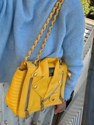 Gul axelväska från Moschino - Cool gul axelväska från Moschino i skinn med detaljer som påminner om en bikerjacka. Väskan har guldfärgade metalldetaljer, kedjerem och quiltad baksida. Perfekt för dig som vill sticka ut med en färgglad och unik accessoar.
