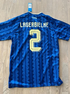 Sverige Adidas matchtröja blå Lagerbielke - Blå Sverige matchtröja från Adidas med gult tryck, nummer 2 och namnet Lagerbielke på ryggen. Tröjan har mönstrade blå ränder, gula detaljer på axlarna och broderat landslagsemblem framtill. Tillverkad i lätt och ventilerande material.