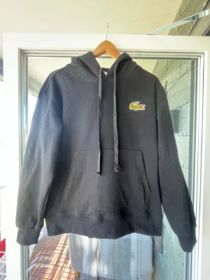 Svart hoodie från Lacoste - Svart hoodie från Lacoste med klassisk krokodillogga på bröstet. Hoodien har huva med dragsko och en stor magficka framtill. Perfekt för en avslappnad och stilren look. Postar den inom max två dagar.