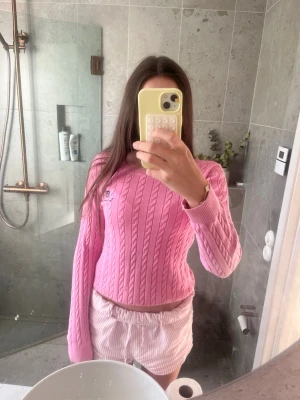 Rosa kabelstickad tröja Bondelid - Snygg rosa kabelstickad tröja från Bondelid med broderad logga och emblem på bröstet. Tröjan har rund halsringning och långa ärmar. Perfekt för en trendig och avslappnad look.