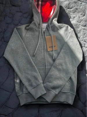 Grå zip hoodie från Burberry - Snygg grå zip hoodie från Burberry med klassisk huva och dragsko. Insidan av huvan har Burberrys ikoniska rutmönster och röd detalj. Tillverkad i mjukt material med ribbade muddar och fickor framtill. Perfekt för en chill och stilren look.