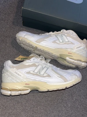 New Balance 1906D vita sneakers - Snygga New Balance 1906D sneakers i vitt och beige med mesh och läderdetaljer. Skorna har en chunky sula med N-ergy dämpning och klassisk N-logga på sidan. Perfekta för dig som gillar retrovibbar och streetwear.