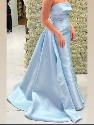 Ljusblå axelbandslös balklänning - Elegant ljusblå balklänning med axelbandslös design och figurnära överdel. Kjolen är vid och golvlång med ett vackert fall. Midjan pryds av ett smalt bälte med glittrande detaljer. Klänningen är tillverkad i ett glansigt, satinliknande material.