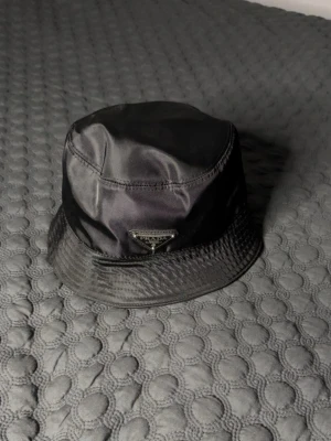 Svart bucket hat från Prada - Snygg svart bucket hat från Prada med den ikoniska metall-loggan framtill. Hatten är tillverkad i ett glansigt syntetmaterial som ger en modern och clean look. Perfekt accessoar för att lyfta din outfit. NYPRIS 5K+