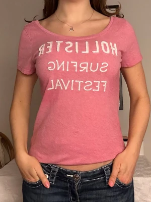 Rosa Hollister top med vitt tryck💗 - Säljer en rosa t-shirt från Hollister med vit text på bröstet. T-shirten har rund halsringning och korta ärmar. Materialet känns mjukt och skönt, troligen bomull. Perfekt för en avslappnad och cool stil. På tredje bilden ser man hur större delen av tröjan är lite blekare än vissa mindre delar💗