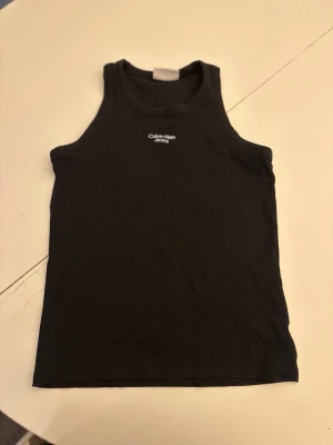 Svart ribbad linnetopp Calvin Klein - Säljer en svart ribbad linnetopp från Calvin Klein Jeans i storlek XS. Toppen har broderad logga framtill och är ärmlös med klassisk rund hals. Perfekt att styla med jeans eller kjol för en clean look.
