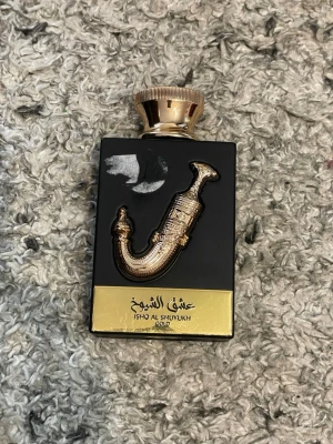 Ishq Al Shuyukh Gold parfym - En lyxig parfymflaska i svart och guld med en dekorativ guldfärgad detalj på framsidan. Flaskan har en elegant, rundad guldkork och en exklusiv design som sticker ut i parfymhyllan. Märket och namnet är tydligt tryckt på en guldfärgad platta längst ner.