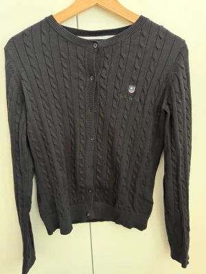 Bondelid - svart kabelstickad kofta Gant - Svart  kabelstickad kofta från Gant med rund halsringning och knappstängning framtill. Liten broderad logga på bröstet och ribbade muddar vid ärmslut och nederkant. Perfekt för lager på lager och klassisk stil.