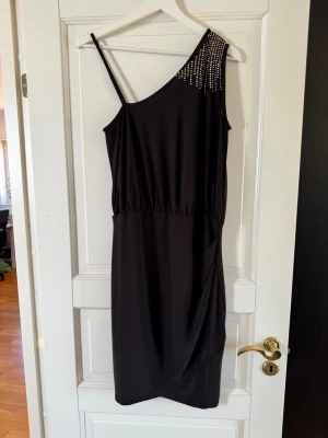 Svart oneshoulder klänning med paljetter - Snygg svart oneshoulder klänning med smal axelrem och paljett-detaljer på axeln. Klänningen har en draperad effekt framtill och resår i midjan för en smickrande passform. Perfekt för dig som vill sticka ut lite extra.