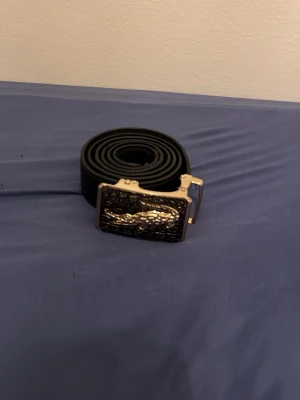 Svart bälte med silverdetalj lacoste  - Snyggt svart bälte med en unik silverspänne som har ett upphöjt mönster. Bältet är stilrent och passar till många olika outfits. Tillverkat i skinn för en klassisk look.